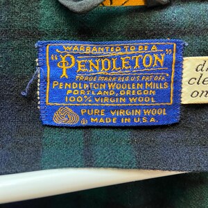 Op de afbeelding: Close-up van een blauw en goud Pendleton-label genaaid op een groen en zwart geruite stof. Het label luidt "Warranted to be a Pendleton" en "Pure Virgin Wool Made in U.S.A."