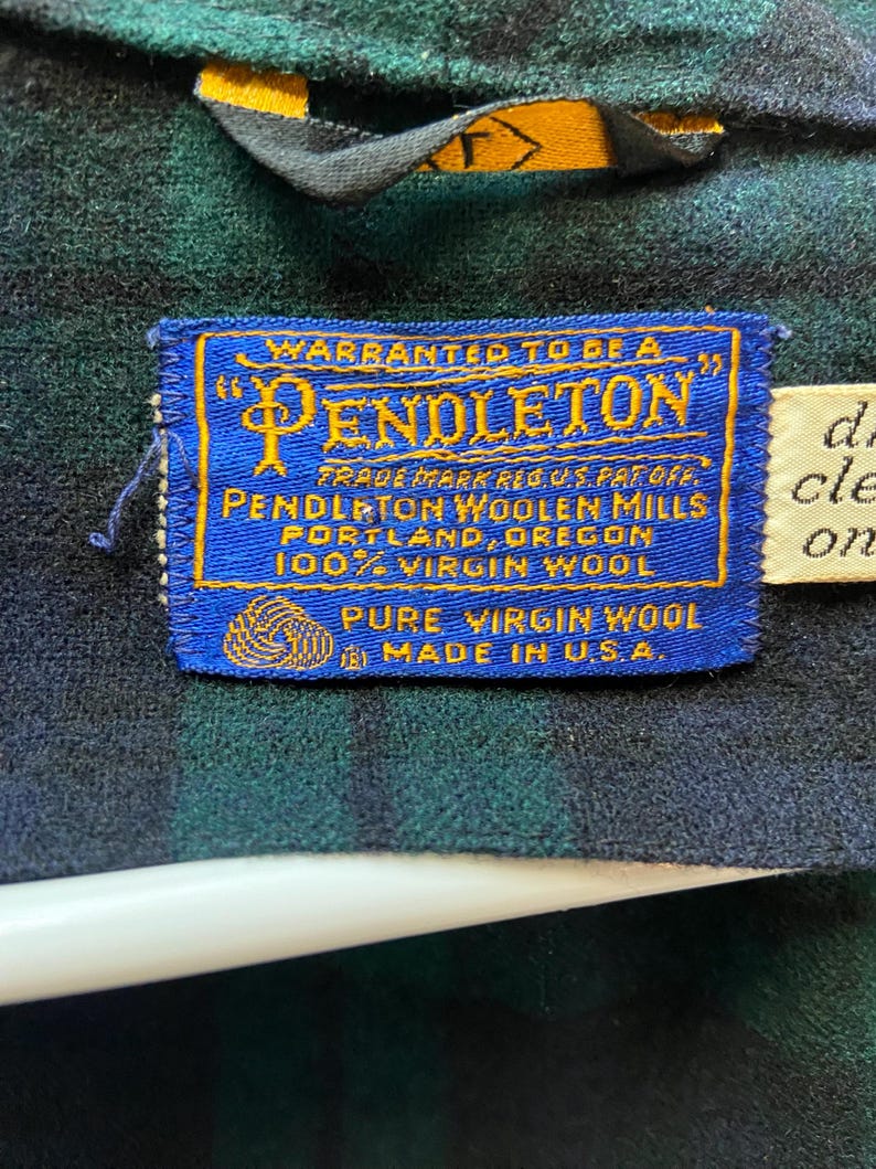 Op de afbeelding: Close-up van een blauw en goud Pendleton-label genaaid op een groen en zwart geruite stof. Het label luidt "Warranted to be a Pendleton" en "Pure Virgin Wool Made in U.S.A."