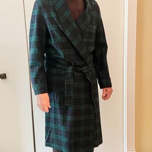 Op de afbeelding: Een lange, geruite badjas in zwart, groen en blauw. De badjas heeft een sjaalkraag en een bijpassende riem. De persoon draagt een zwarte broek en een bruin overhemd. De badjas is een klassieke stijl.