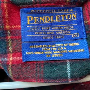 Op de afbeelding: Close-up van een blauw Pendleton-label op een rood, groen en geel geruit overhemd. Het label luidt "WARRANTED TO BE A PENDLETON 100% PURE VIRGIN WOOL PORTLAND, OREGON SINCE 1863." Het overhemd is XL en geassembleerd in Mexico.