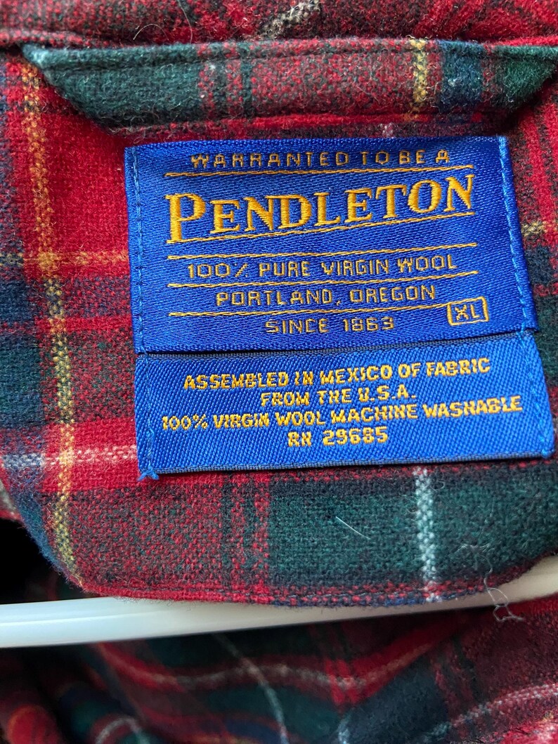 Op de afbeelding: Close-up van een blauw Pendleton-label op een rood, groen en geel geruit overhemd. Het label luidt "WARRANTED TO BE A PENDLETON 100% PURE VIRGIN WOOL PORTLAND, OREGON SINCE 1863." Het overhemd is XL en geassembleerd in Mexico.