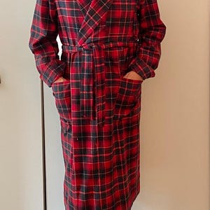 Op de afbeelding: Een lange, rood-zwarte geruite badjas met een sjaalkraag en een bijpassende riem. De badjas heeft twee voorzakken en wordt gedragen over een donker overhemd en een zwarte broek. De badjas is gemaakt van een zacht, warm materiaal.