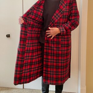 Op de afbeelding: Een lange, rood-zwarte geruite badjas met een sjaalkraag. De badjas is open en onthult een donker overhemd en broek. De badjas is gemaakt van een zacht, warm materiaal.