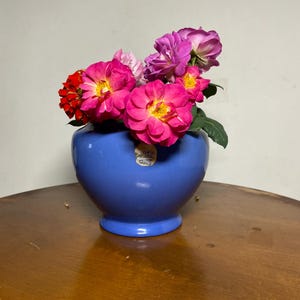May include: A glossy blue ceramic vase with a rounded body and a flared base, containing a bouquet of vibrant pink, red, and purple roses. A small, circular gold-toned sticker with illegible text is affixed to the front of the vase.