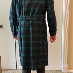 Op de afbeelding: Een lange, geruite badjas in groen- en marineblauwtinten. De badjas heeft een bijpassende riem en lange mouwen. De persoon op de foto draagt een zwarte broek en zwarte schoenen. De badjas is gemaakt van een zacht, mogelijk flanellen materiaal.