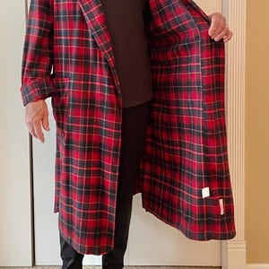 Op de afbeelding: Een lange, rood-zwarte geruite badjas met een sjaalkraag. De badjas is open en onthult een donkerbruin shirt en een zwarte broek. De binnenkant van de badjas is ook rood-zwart geruit. Zwarte sokken zijn zichtbaar.