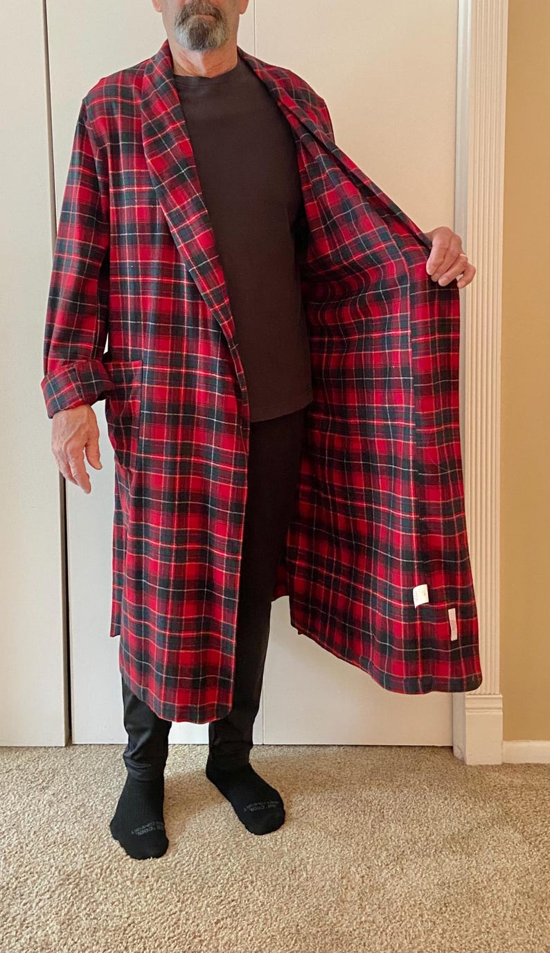 Op de afbeelding: Een lange, rood-zwarte geruite badjas met een sjaalkraag. De badjas is open en onthult een donkerbruin shirt en een zwarte broek. De binnenkant van de badjas is ook rood-zwart geruit. Zwarte sokken zijn zichtbaar.