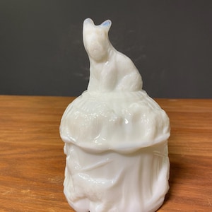 May include: A white, opalescent glass trinket box with a cat figurine on top. The cat is sitting atop a textured lid, and the box has a draped fabric design. The box is made of a glossy, opaque glass.
