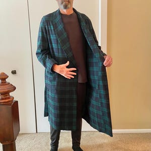 Op de afbeelding: Een lange, geruite badjas in groen en marineblauw. De badjas heeft een klassiek tartanpatroon en een sjaalkraag. De persoon draagt een bruin shirt en een zwarte broek. De badjas is gemaakt van een zacht, comfortabel materiaal.