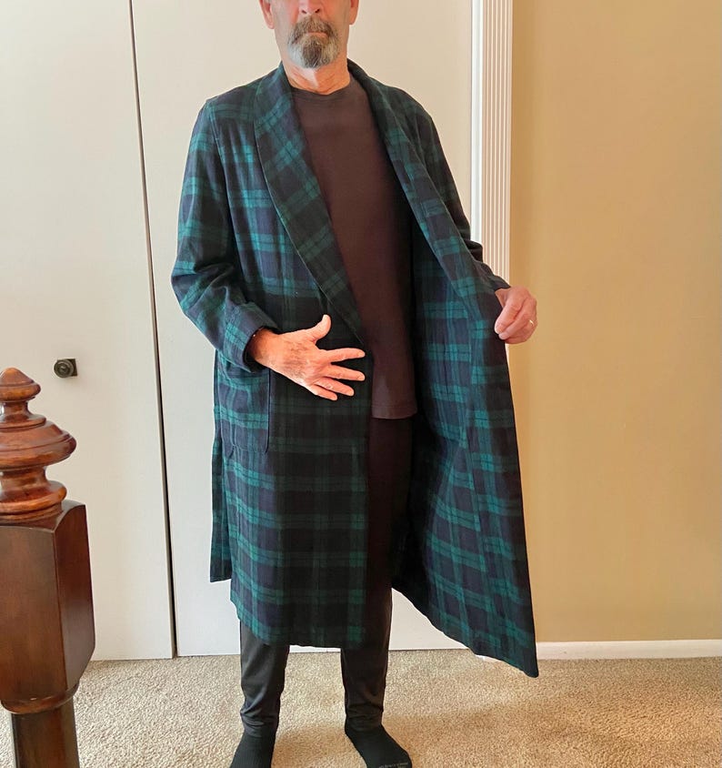 Op de afbeelding: Een lange, geruite badjas in groen en marineblauw. De badjas heeft een klassiek tartanpatroon en een sjaalkraag. De persoon draagt een bruin shirt en een zwarte broek. De badjas is gemaakt van een zacht, comfortabel materiaal.