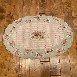 May include: An oval rug featuring a central floral bouquet surrounded by a pink and white diamond lattice pattern. The scalloped border is light green with a repeating pattern of pink roses and green leaves throughout the design.