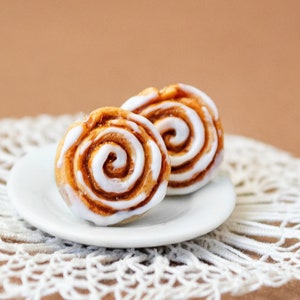 Könnte beinhalten: Zwei Miniatur-Zimtschnecken mit weißem Zuckerguss auf einem kleinen weißen Teller. Die Brötchen haben ein spiralförmiges Design und liegen auf einer weißen Spitzendecke. Der Hintergrund ist einfarbig beige.