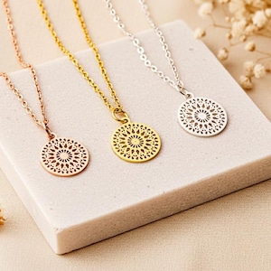 Könnte beinhalten: Drei zarte Halsketten mit runden Anhängern. Eine ist roségoldfarben, eine goldfarben und die andere silberfarben. Jeder Anhänger hat ein filigranes, offenes Design. Die Halsketten werden auf einem weißen, rechteckigen Block präsentiert.