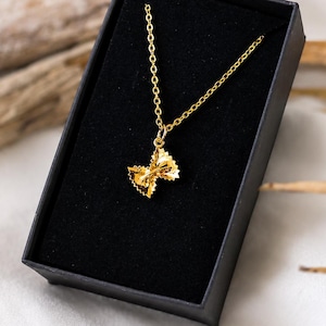 Könnte beinhalten: Eine goldfarbene Halskette mit einem kleinen, strukturierten Anhänger in Form einer Schleifennudel, der an einer feinen Gliederkette hängt und in einer rechteckigen schwarzen Schachtel mit schwarzem Samtfutter präsentiert wird.