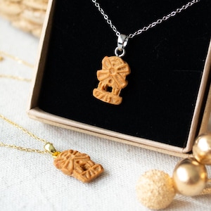 Könnte beinhalten: Zwei Halsketten mit Windmühlen-Anhängern. Eine Halskette hat eine silberne Kette, die andere eine goldene Kette. Die braunen Windmühlen-Anhänger werden in einer kleinen Schachtel mit schwarzem Innenraum präsentiert. Der Hintergrund umfasst einen geflochtenen Korb und dekorative goldene Kugeln.