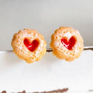 Può includere: Due biscotti alla marmellata a forma di cuore spolverati di zucchero a velo. I biscotti hanno un bordo smerlato e un ripieno rosso brillante a forma di cuore. I biscotti sono di colore marrone dorato chiaro.