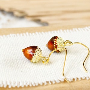 Könnte beinhalten: Ein Paar goldene Eichel-Ohrringe mit braunen und goldenen Akzenten. Die Ohrringe sind im Hänge-Stil mit einem kleinen Eichel-Anhänger.