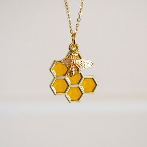 Può includere: Collana d'oro con un ciondolo a forma di favo e un piccolo ciondolo a forma di ape. Il favo è giallo e l'ape è dorata.