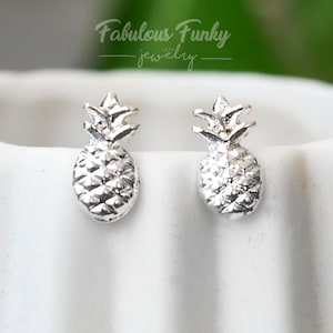 Könnte beinhalten: Ein Paar silberfarbene Ohrstecker in Form von Ananas. Die Ohrringe weisen detaillierte, strukturierte Oberflächen auf, die die Schale und die Krone der Ananas darstellen. Im Hintergrund ist der Text "Fabulous Funky jewelry" zu sehen.