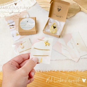 K&ouml;nnte beinhalten: Eine Kollektion goldfarbener Schmuckst&uuml;cke, darunter Halsketten mit Hunde- und Tropfenanh&auml;ngern, pr&auml;sentiert in braunen Pappschachteln und auf Marken-Pr&auml;sentationskarten mit dem Text Fabulous Funky Jewelry und Mit Liebe Verpackt.