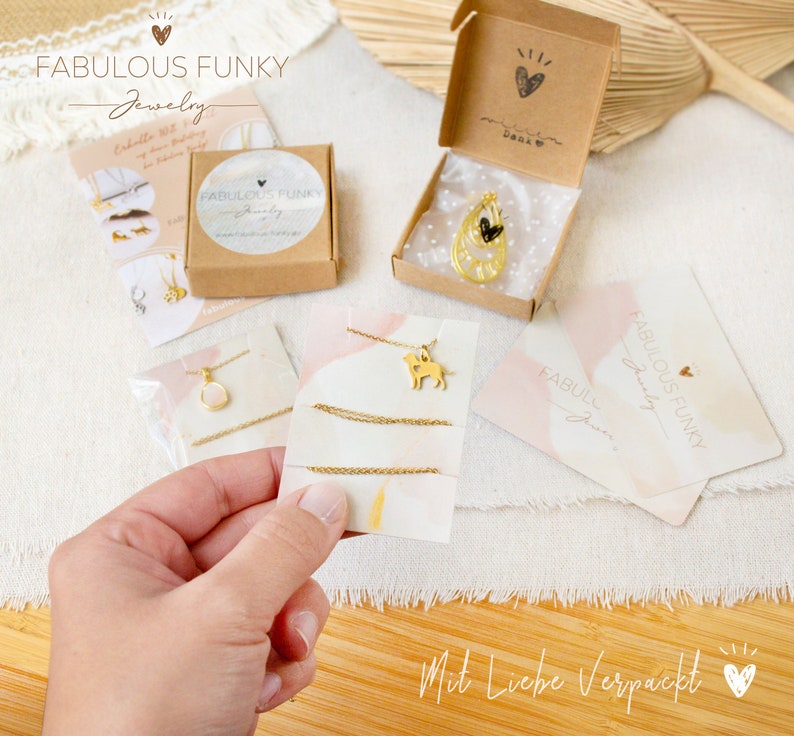 K&ouml;nnte beinhalten: Eine Kollektion goldfarbener Schmuckst&uuml;cke, darunter Halsketten mit Hunde- und Tropfenanh&auml;ngern, pr&auml;sentiert in braunen Pappschachteln und auf Marken-Pr&auml;sentationskarten mit dem Text Fabulous Funky Jewelry und Mit Liebe Verpackt.