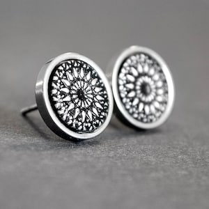 Op de afbeelding: Een paar zilverkleurige oorstekers. Elke oorbel heeft een cirkelvormig ontwerp met een zwart-wit bloemenpatroon. De oorbellen staan tegen een grijze achtergrond.