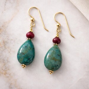 May include: A pair of gold-colored drop earrings. Each earring features a teardrop-shaped turquoise stone, a small red bead, and gold-colored accents. The earrings have a simple hook design.