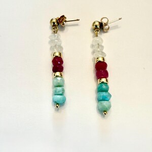 May include: A pair of dangle earrings with gold-colored posts and accents. Each earring features a vertical arrangement of white, red, and turquoise-colored gemstone beads. The earrings are displayed against a white background.