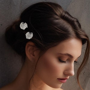 May include: A woman wearing a white off-the-shoulder gown with a simple, elegant design. She has her hair pulled back and is wearing two small, delicate floral hair clips.