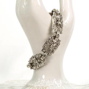 May include: A silver-toned link bracelet featuring a geometric design with embedded clear rhinestones. The bracelet consists of rectangular segments connected by smaller links, creating a structured appearance with a metallic finish and sparkling stones.
