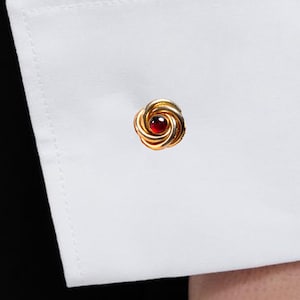 May include: A gold-toned metal cufflink featuring a swirling, circular design with a small, round, deep red gemstone set in the center. The cufflink is fastened through the buttonhole of a crisp, white, long-sleeved dress shirt cuff.