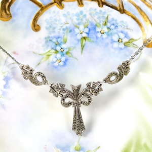 Puede incluir: Un collar de tono plateado con un colgante central en forma de lazo y una ca&iacute;da vertical, todo adornado con peque&ntilde;as piedras oscuras facetadas. La cadena consta de eslabones delicados que se conectan a la pieza central ornamentada y sim&eacute;trica.