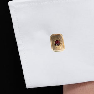 May include: A crisp white dress shirt sleeve featuring a double French cuff secured by a rectangular gold-toned cufflink. The cufflink is centered with a small, deep red circular gemstone. The shirt fabric is smooth, opaque, and bright white.