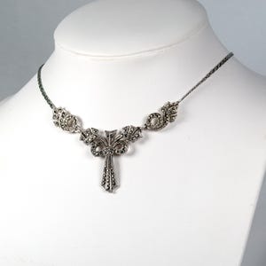 Puede incluir: Un collar de tono plateado con un colgante central en forma de lazo y una ca&iacute;da vertical, adornado con numerosas piedras de marcasita oscuras y facetadas. La cadena es de eslabones met&aacute;licos finos y flexibles con detalles laterales a juego.