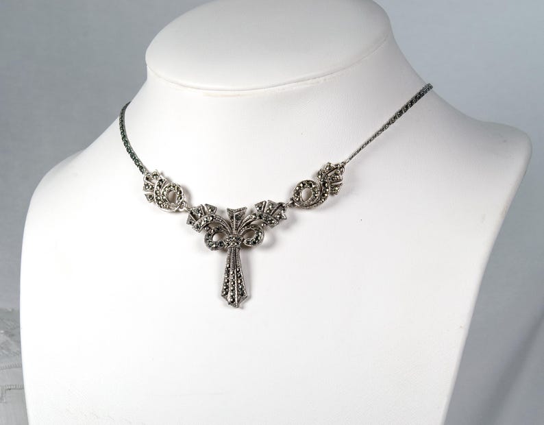 Puede incluir: Un collar de tono plateado con un colgante central en forma de lazo y una ca&iacute;da vertical, adornado con numerosas piedras de marcasita oscuras y facetadas. La cadena es de eslabones met&aacute;licos finos y flexibles con detalles laterales a juego.