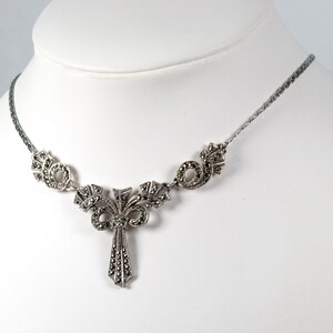 Puede incluir: Un collar de tono plateado con un colgante central que presenta una ca&iacute;da vertical en forma de abanico y adornos sim&eacute;tricos en espiral. Toda la pieza est&aacute; incrustada con peque&ntilde;as piedras oscuras facetadas y unida a una cadena fina.