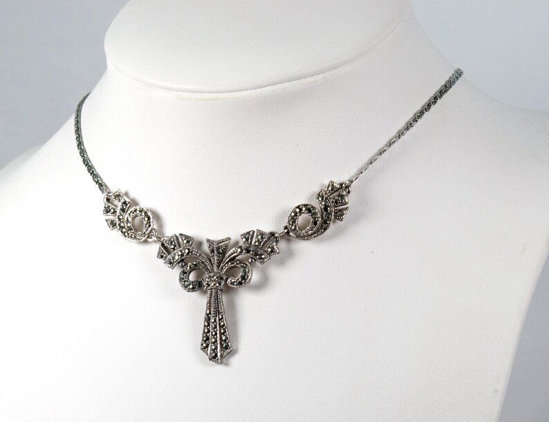 Puede incluir: Un collar de tono plateado con un colgante central que presenta una ca&iacute;da vertical en forma de abanico y adornos sim&eacute;tricos en espiral. Toda la pieza est&aacute; incrustada con peque&ntilde;as piedras oscuras facetadas y unida a una cadena fina.