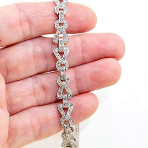 May include: A silver bracelet with a series of linked, bow-shaped designs, each encrusted with small, clear gemstones. The bracelet is held against a white background, showcasing its intricate details and sparkling appearance.