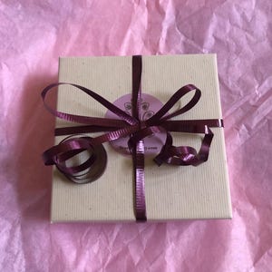 Puede incluir: Una caja de regalo cuadrada de cart&oacute;n beige claro atada con una cinta met&aacute;lica de color morado oscuro. Una pegatina circular de color morado claro con un dise&ntilde;o gr&aacute;fico oscuro y el texto TrueJoy.com est&aacute; centrada en la tapa, bajo el nudo.