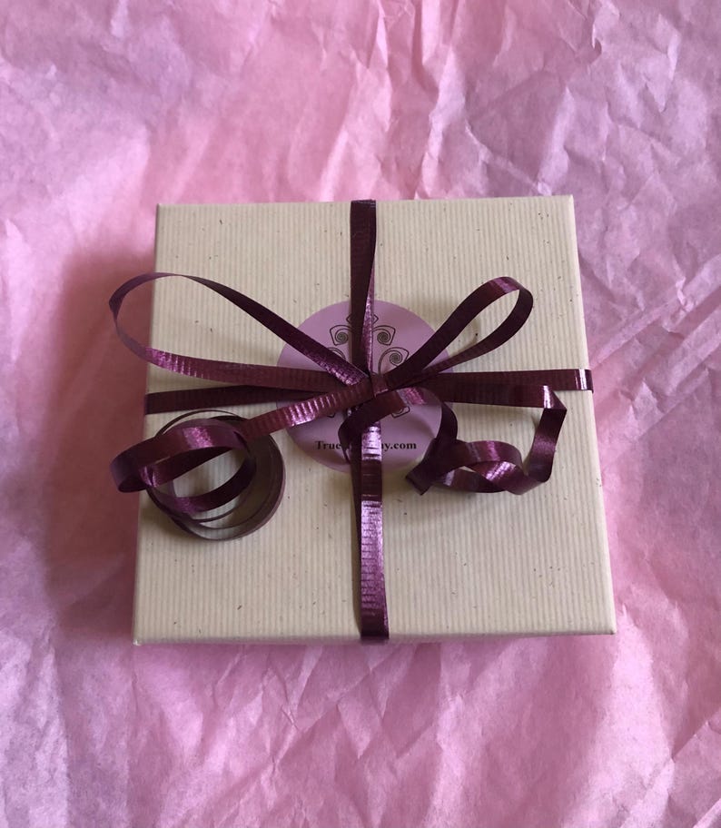 Puede incluir: Una caja de regalo cuadrada de cart&oacute;n beige claro atada con una cinta met&aacute;lica de color morado oscuro. Una pegatina circular de color morado claro con un dise&ntilde;o gr&aacute;fico oscuro y el texto TrueJoy.com est&aacute; centrada en la tapa, bajo el nudo.