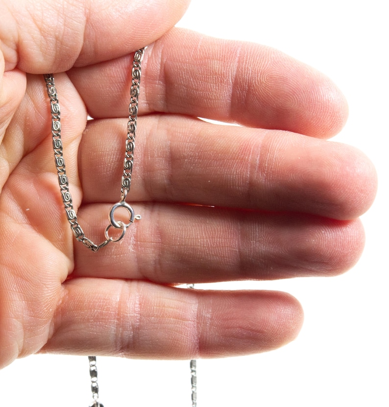 Puede incluir: Un collar de cadena de metal plateado que presenta un patr&oacute;n repetitivo de eslabones intrincados en forma de volutas u ondas. La cadena est&aacute; asegurada con un mecanismo de cierre de resorte circular, mostrando un acabado met&aacute;lico pulido.