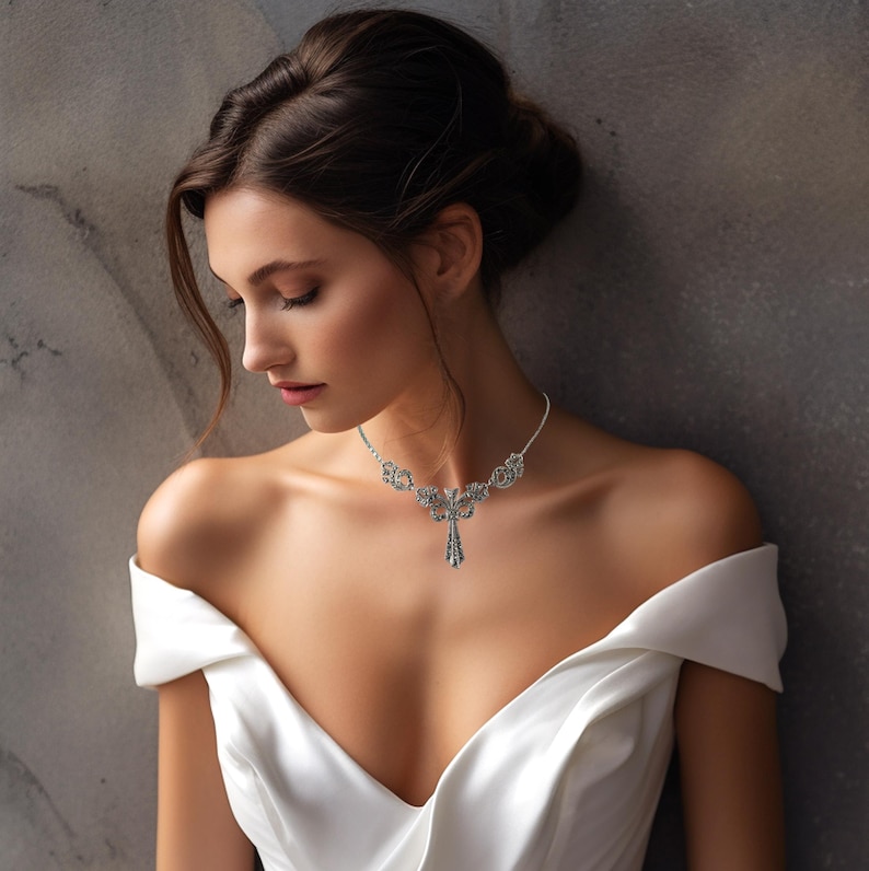 Puede incluir: Un collar de tono plateado que presenta un colgante central ornamentado con intrincados detalles de filigrana y peque&ntilde;as piedras transparentes, suspendido de una cadena delicada que descansa sobre la clav&iacute;cula, con una prenda blanca de hombros descubiertos.