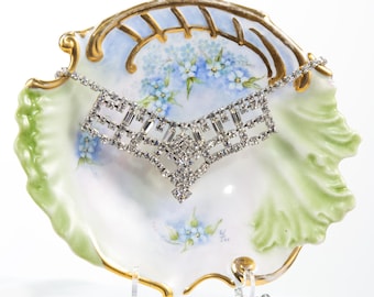 May include: A silver rhinestone necklace with a geometric design, displayed on a white porcelain dish with blue and green floral accents.