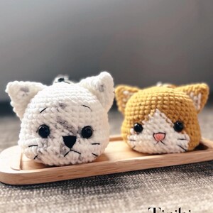 May include: Two handmade crochet cat head ornaments. One is white with grey markings, the other is orange and white. Both have black eyes and pink noses. They sit on a small wooden tray. The text "Tinibi" and website address are at the bottom.