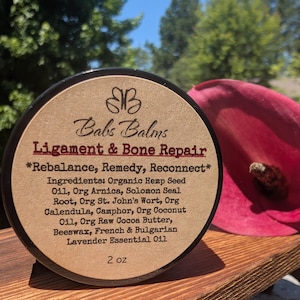 May include: A round container of Babs Balms Ligament & Bone Repair balm with a tan label. The label lists ingredients including organic hemp seed oil, arnica, Solomon seal root, St. John's wort, calendula, camphor, coconut oil, cocoa butter, and beeswax. 2 oz.
