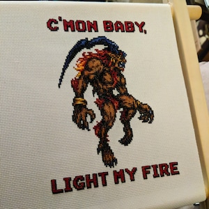 May include: Cross-stitch embroidery of a red, orange, and yellow fire demon with a blue tail, standing on a white background. The text "C'mon baby, light my fire" is stitched in red below the demon.