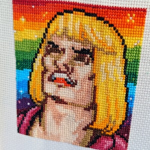 May include: Cross-stitch embroidery of a person with long blonde hair, wearing a pink and purple outfit. The background is a rainbow.
