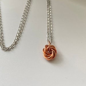 May include: A silver chain necklace with a copper-colored pendant in a woven knot design. The necklace is displayed on a white surface next to a gray box with the text "JCLEECOLLECTION". A floral patterned box is also visible.