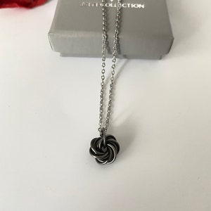 Peut inclure: Un collier en chaîne argentée avec un pendentif en nœud noir. Le collier est dans une petite boîte grise avec le texte "JCB COLLECTION" dessus.