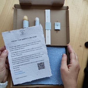Può includere: Una scatola di cartone aperta contenente un kit per quaderno fai-da-te. Il kit include carta azzurra, un ago, strumenti in legno e un foglio di istruzioni con testo italiano e un codice QR.